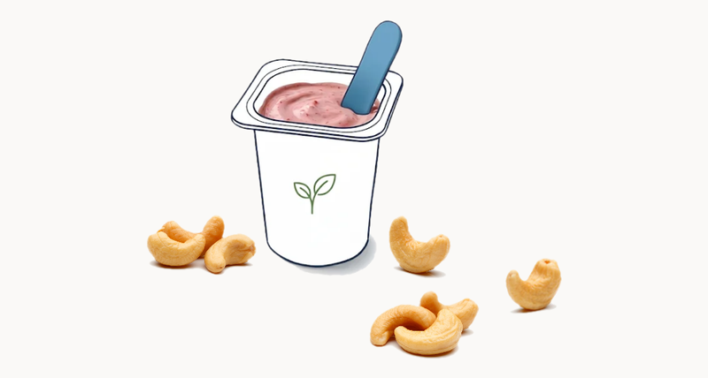 An illustrated cup of cashew-based plant-based yogurt with a pink spoon, a green leaf logo, and whole cashews scattered around the base on a cream background.
